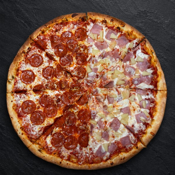 A whole pizza, half pepperoni and half ham and pineapple, on a dark textured surface.