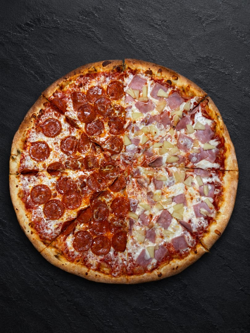 A whole pizza, half pepperoni and half ham and pineapple, on a dark textured surface.