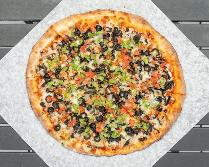 Whole vegetarian pizza with black olives, green peppers, onions, and tomatoes on parchment paper.