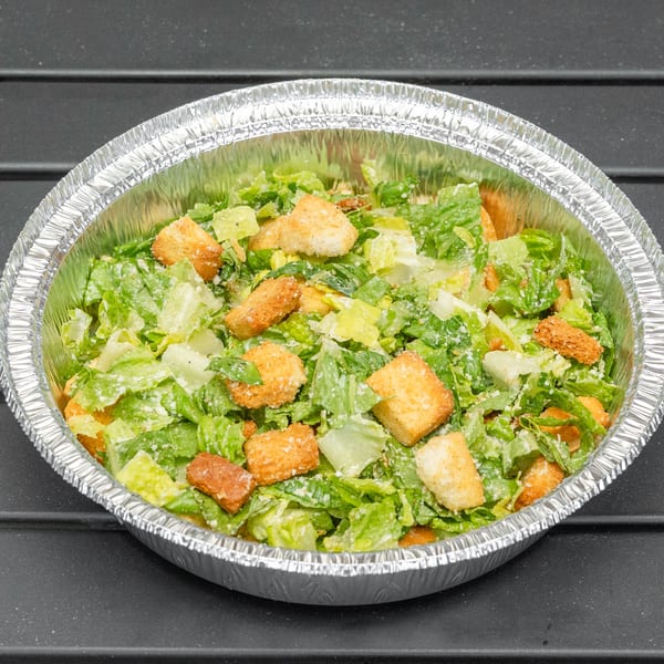 Caesar salad with croutons and dressing in a foil take-out container on a dark surface.