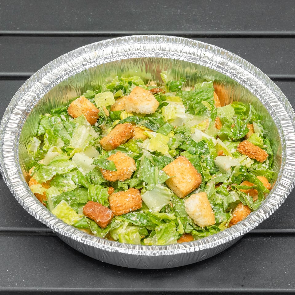 Caesar salad with croutons and dressing in a foil take-out container on a dark surface.