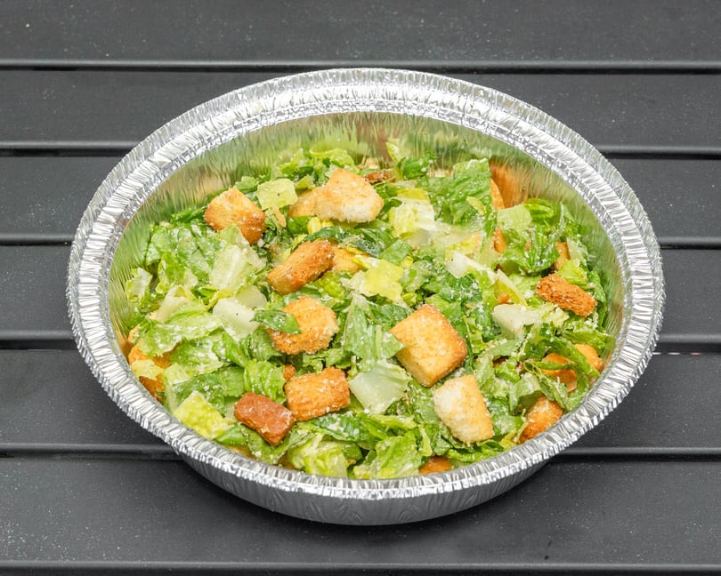 Caesar salad with croutons and dressing in a foil take-out container on a dark surface.
